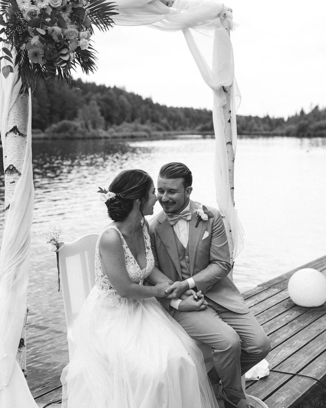 Brautpaar sitzt auf einem Steg, Braut mit weißem Hochzeitskleid, Make-up und gesteckter Brautfrisur. Haare und Make-up von Denise Drahtmueller