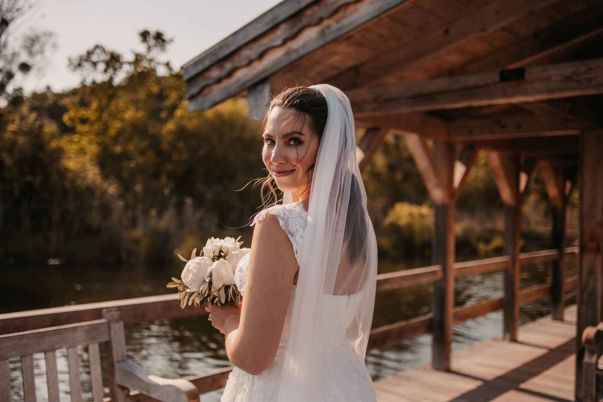 Braut steht auf einem Steg mit Blumenstraß in der Hand. Haare und Make-up von Denise Drahtmueller