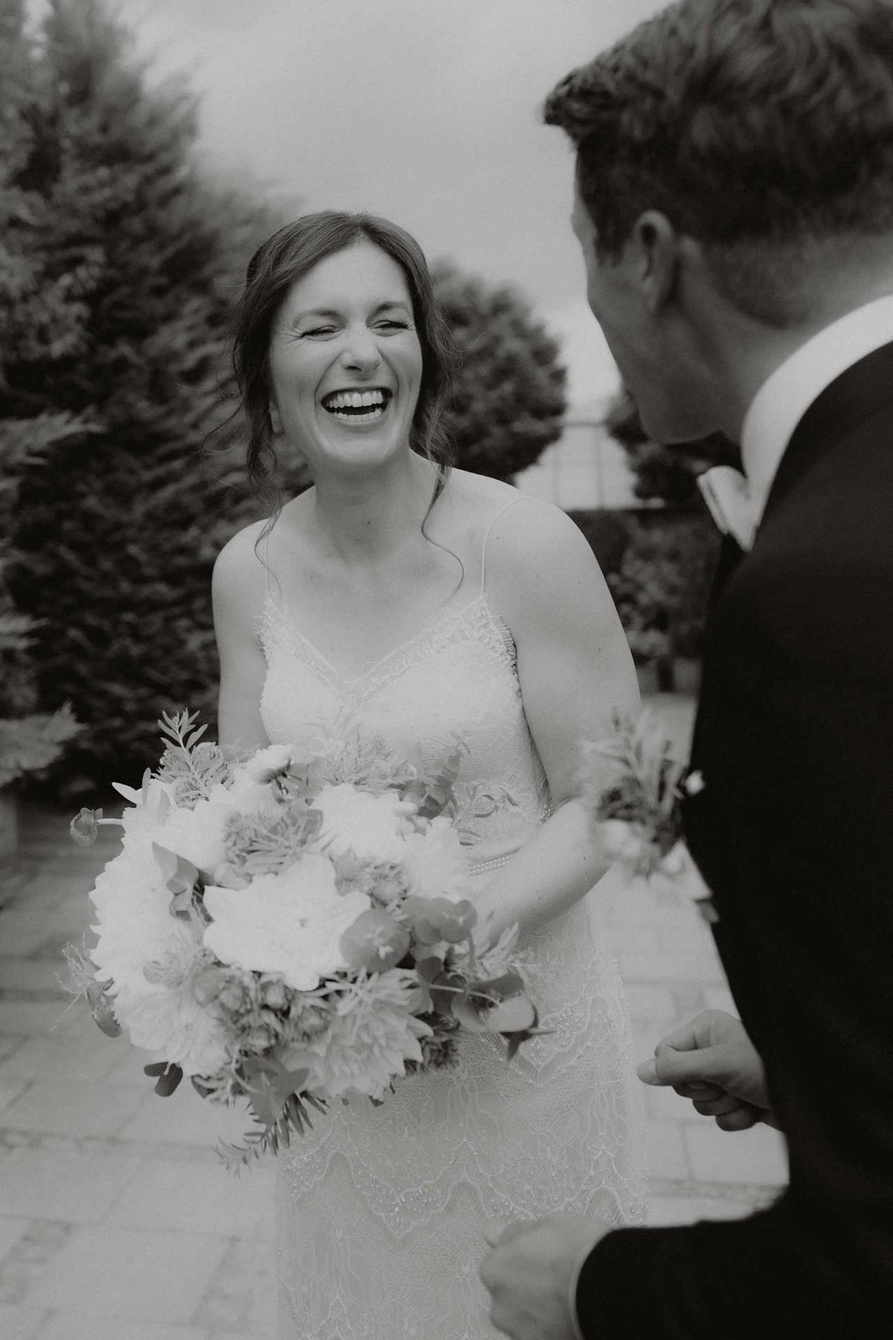 Braut lacht und steht mit Blumenstrauß vor ihrem Bräutigam. Haare und Make-up von Denise Drahtmueller