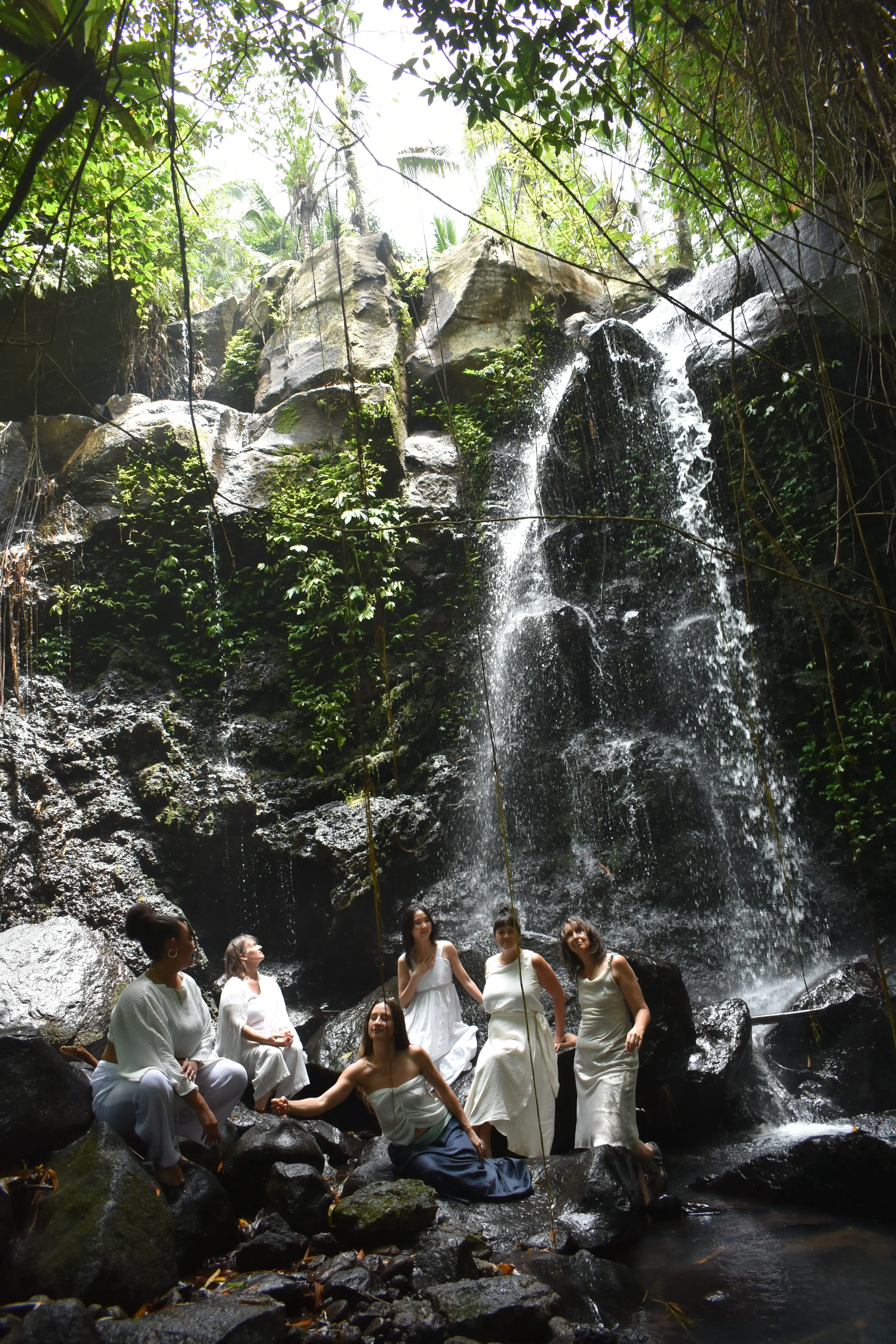 A group of women gathered at the base of a towering, moss-covered waterfall in the Tabanan rainforest during a spiritual wellness retreat.