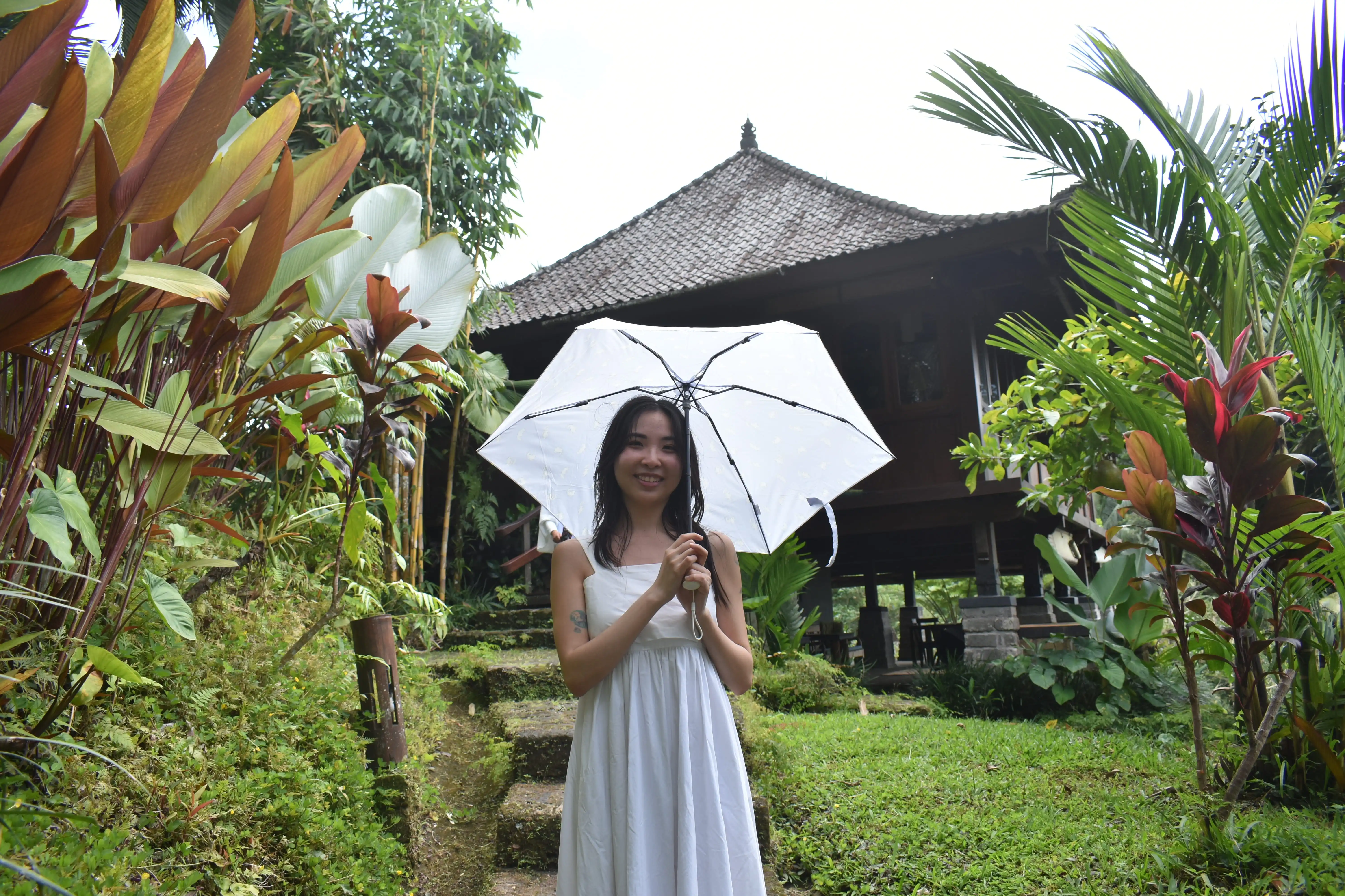 A woman in a white dress holding a white umbrella while walking down stairs toward a traditional thatched-roof bungalow in the lush Tabanan jungle.