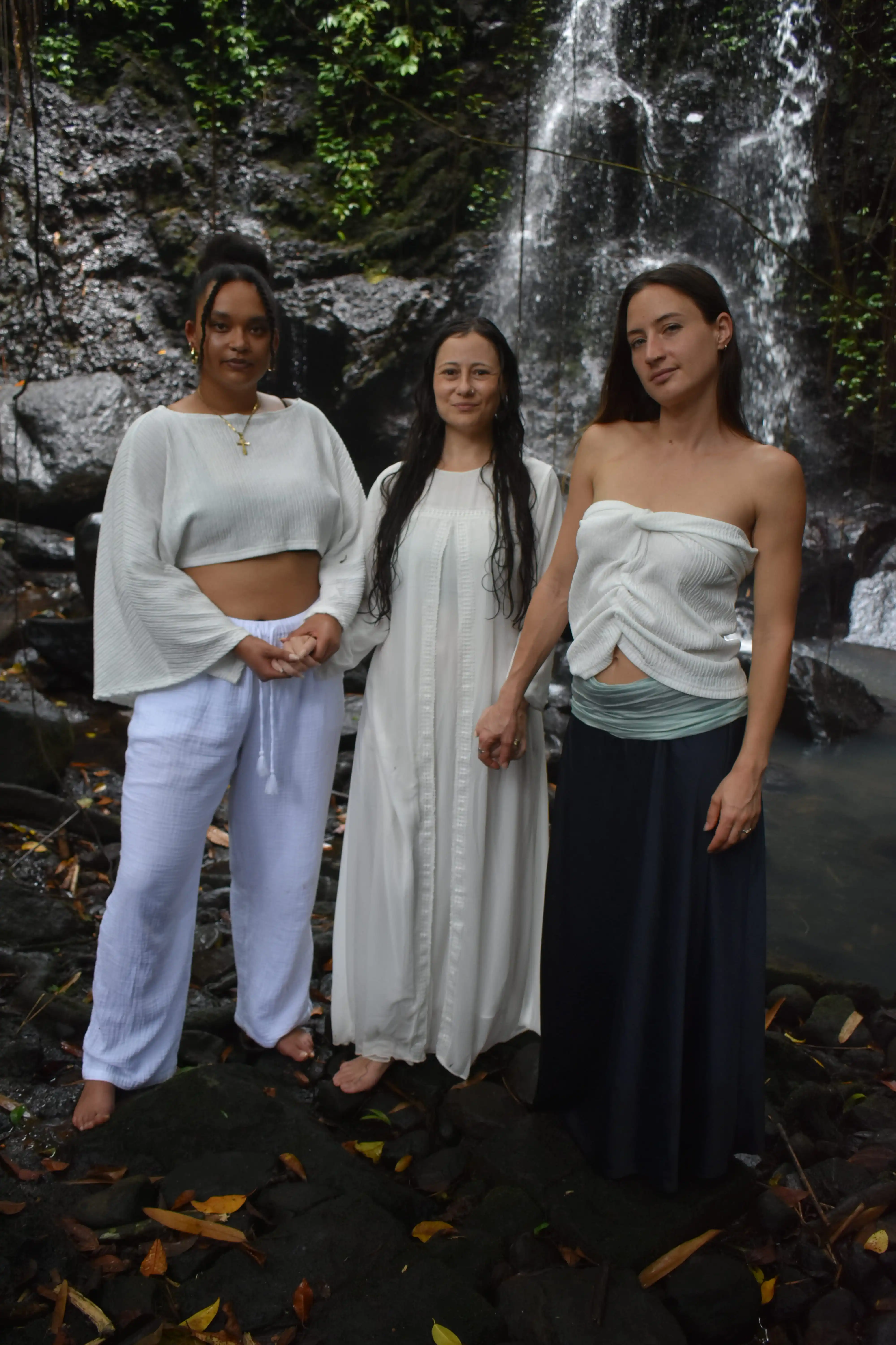 Three women in white clothing holding hands in front of a cascading jungle waterfall in Tabanan, Bali.