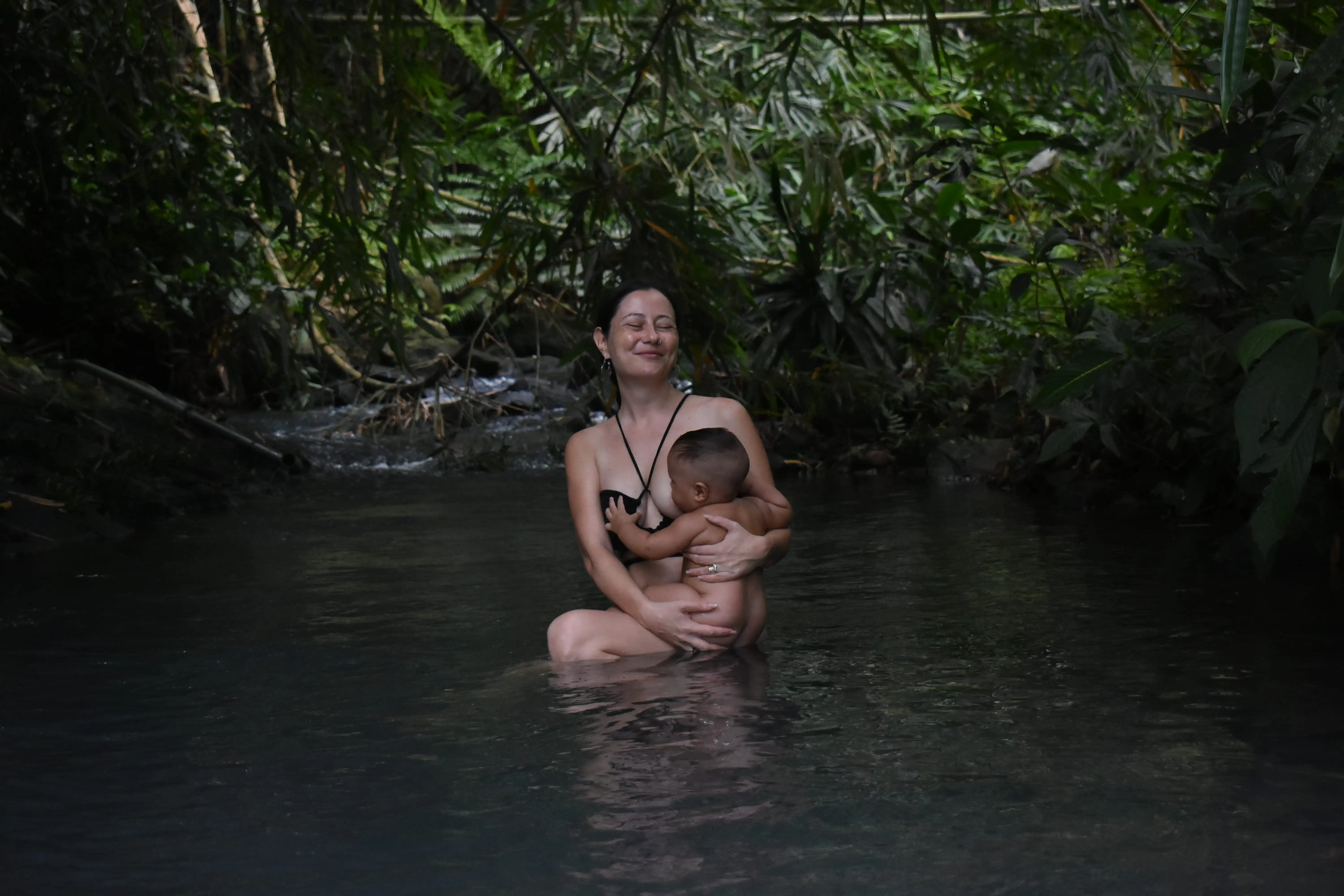 Carla, a psychic and mother, peacefully breastfeeding her young son while sitting in a serene, shaded jungle stream in Tabanan, Bali.