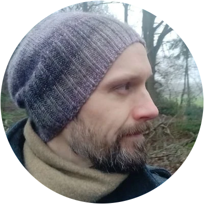 White man with a beard wearing a grey and mauve knitted hat. He has a short beard with some grey in it. Behind are some trees.