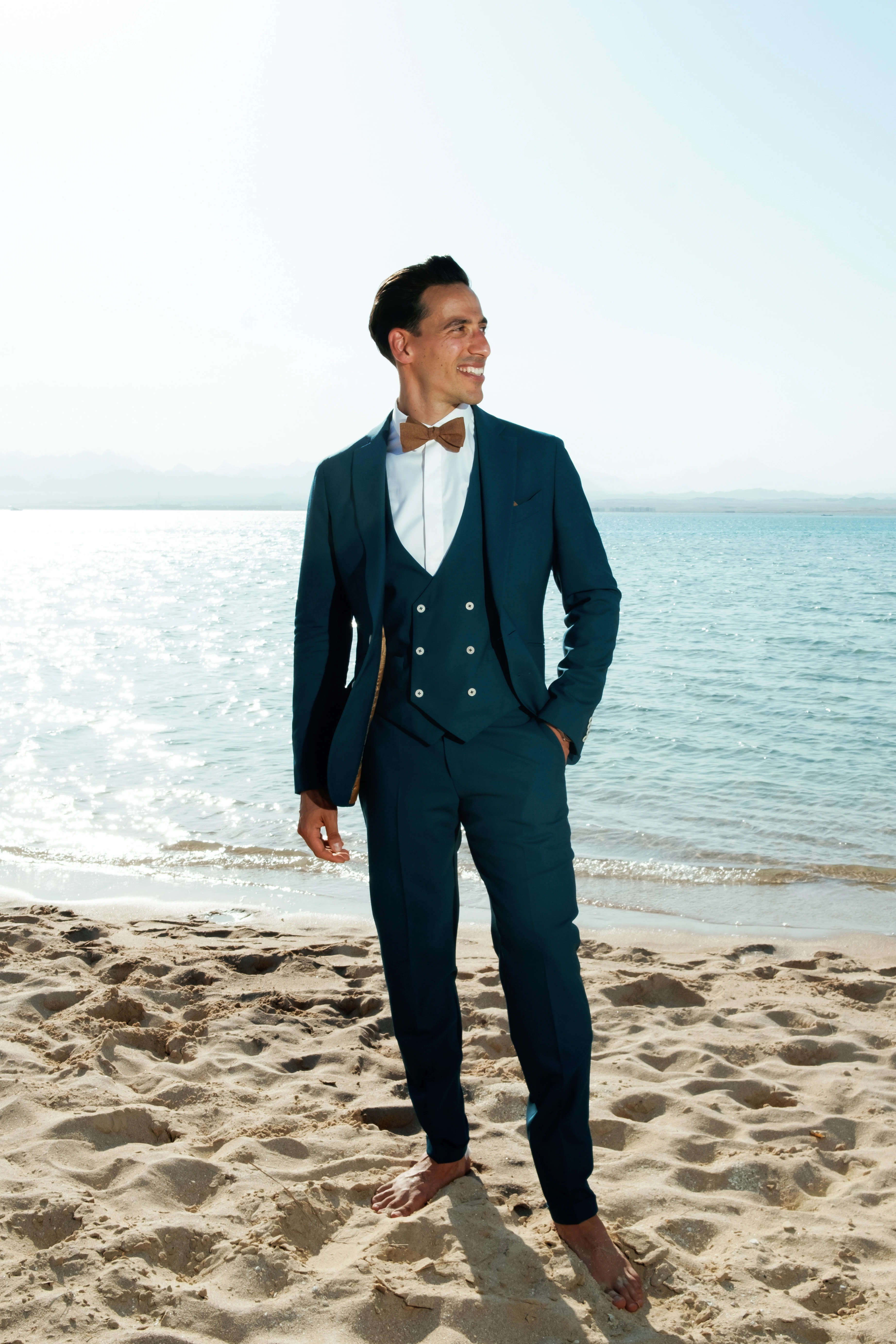 A smiling climber with a helmet and harness seated on a rocky ledge, with a lush green forest and a lake in the background in the Vosges mountains of France.