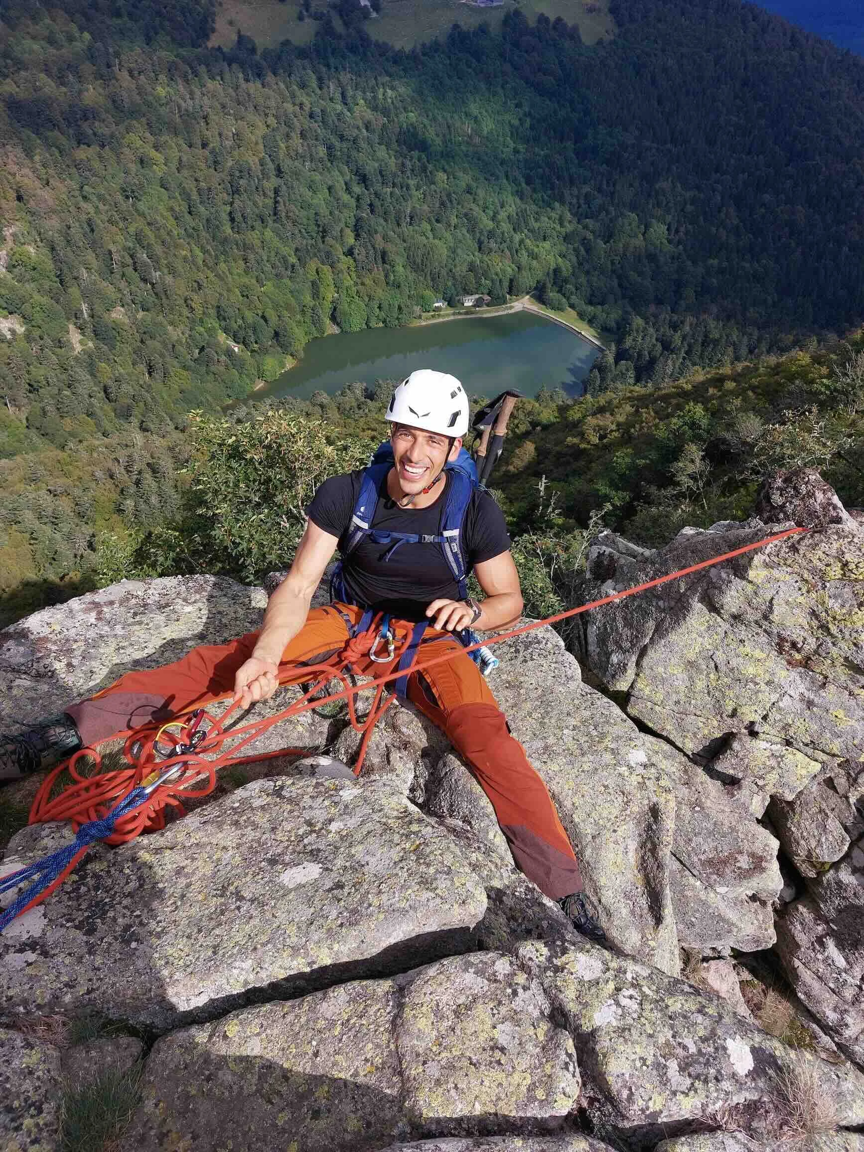 A smiling climber with a helmet and harness seated on a rocky ledge, with a lush green forest and a lake in the background in the Vosges mountains of France.