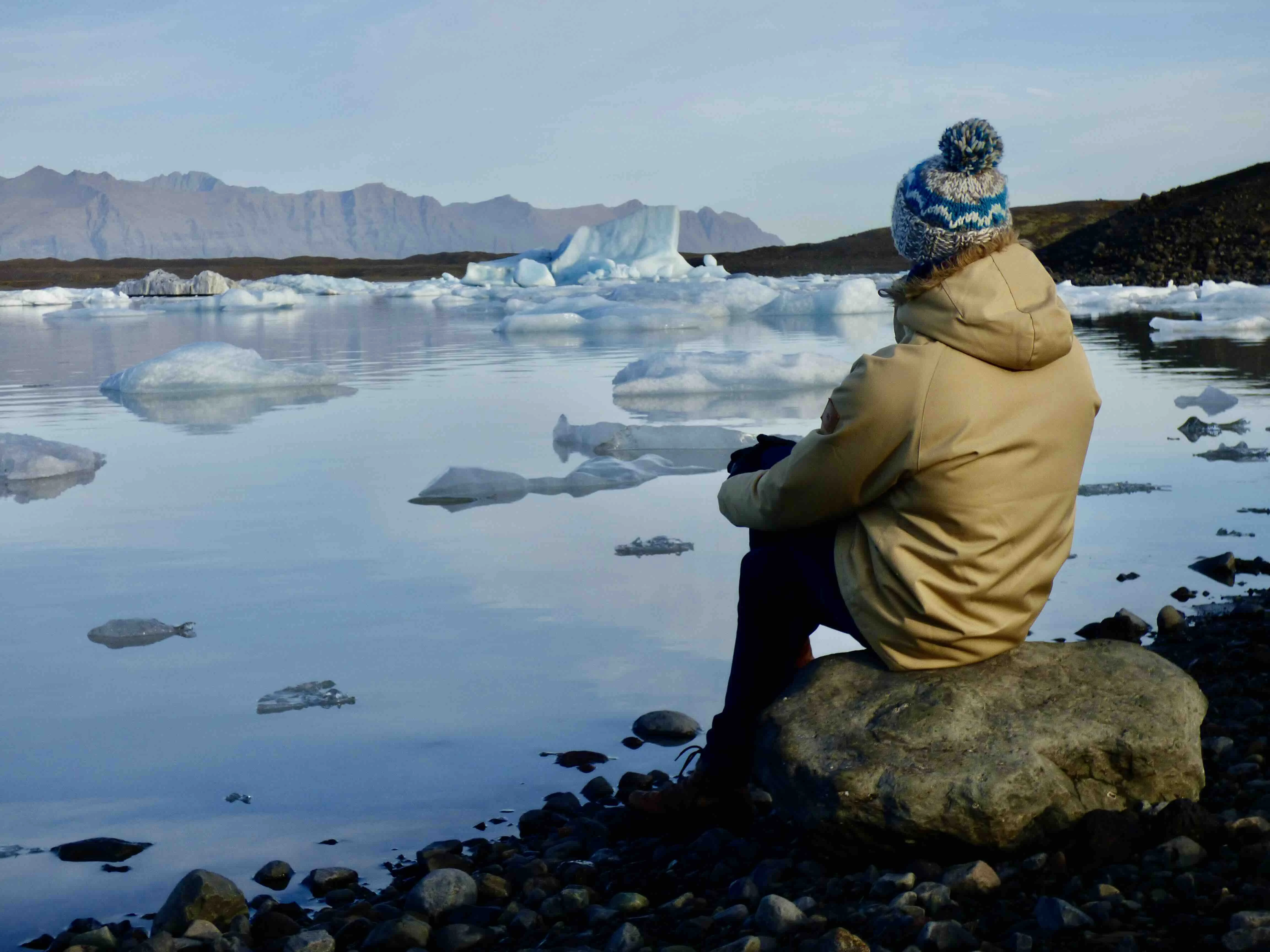 A person seated on a rock wearing a beanie and jacket, gazing at the calm waters with floating icebergs in Iceland.