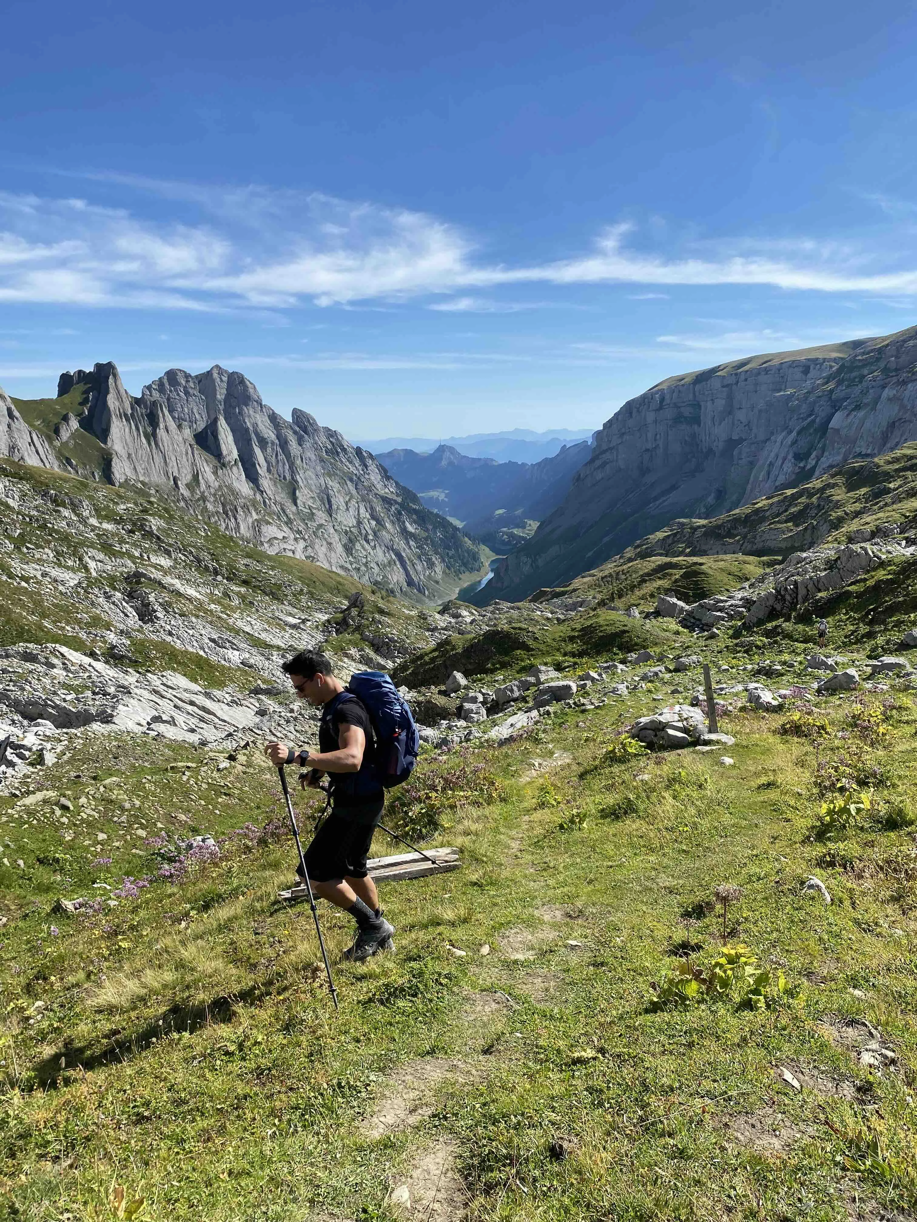 A hiker with a backpack using trekking poles on a mountain trail with scenic views of rocky peaks and a valley in the Appenzell region of the Swiss mountains.